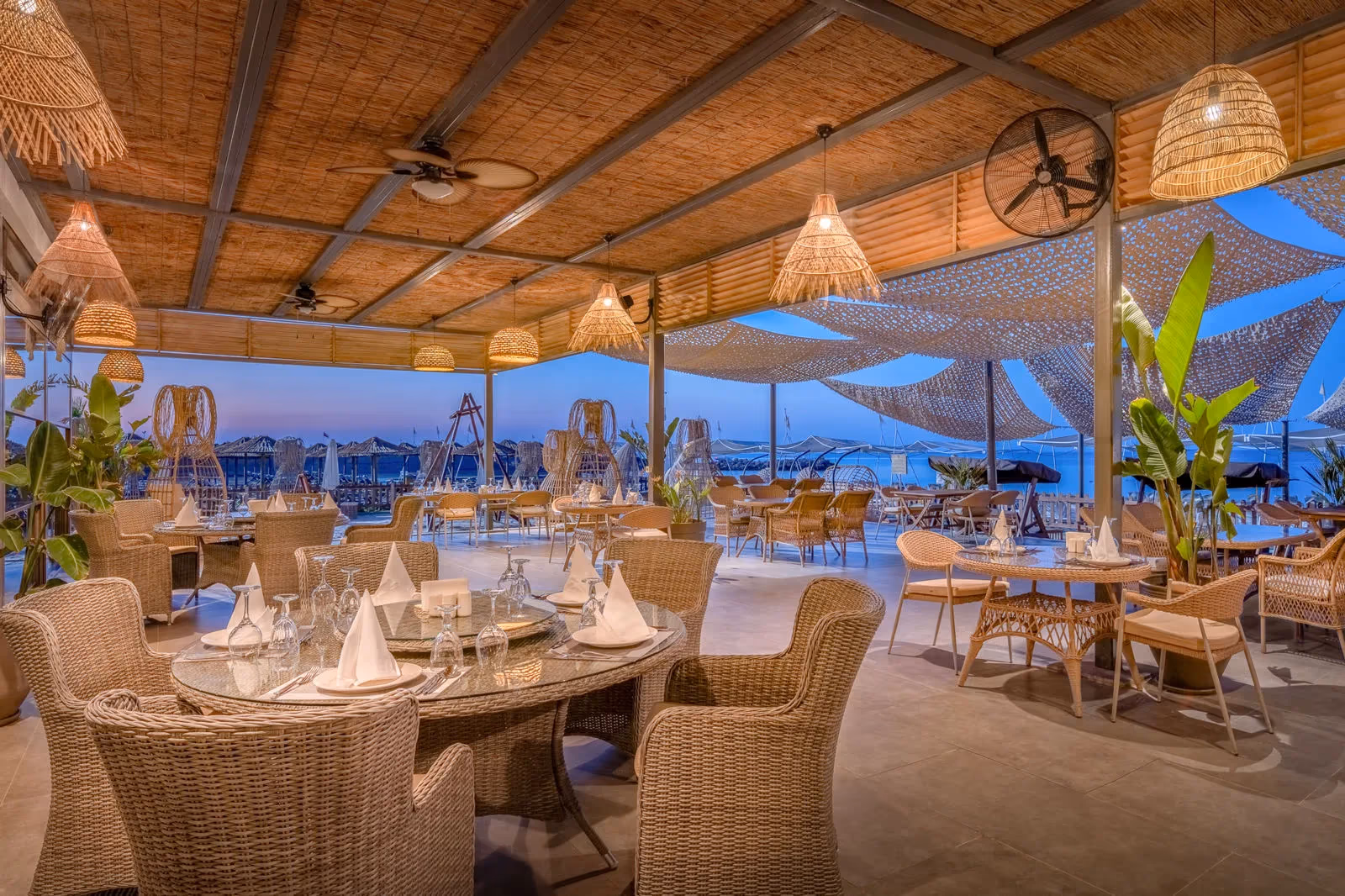 Overdekt restaurantterras met rieten stoelen en gedekte tafels in de avondschemering bij Delphin BE Grand Resort in Lara