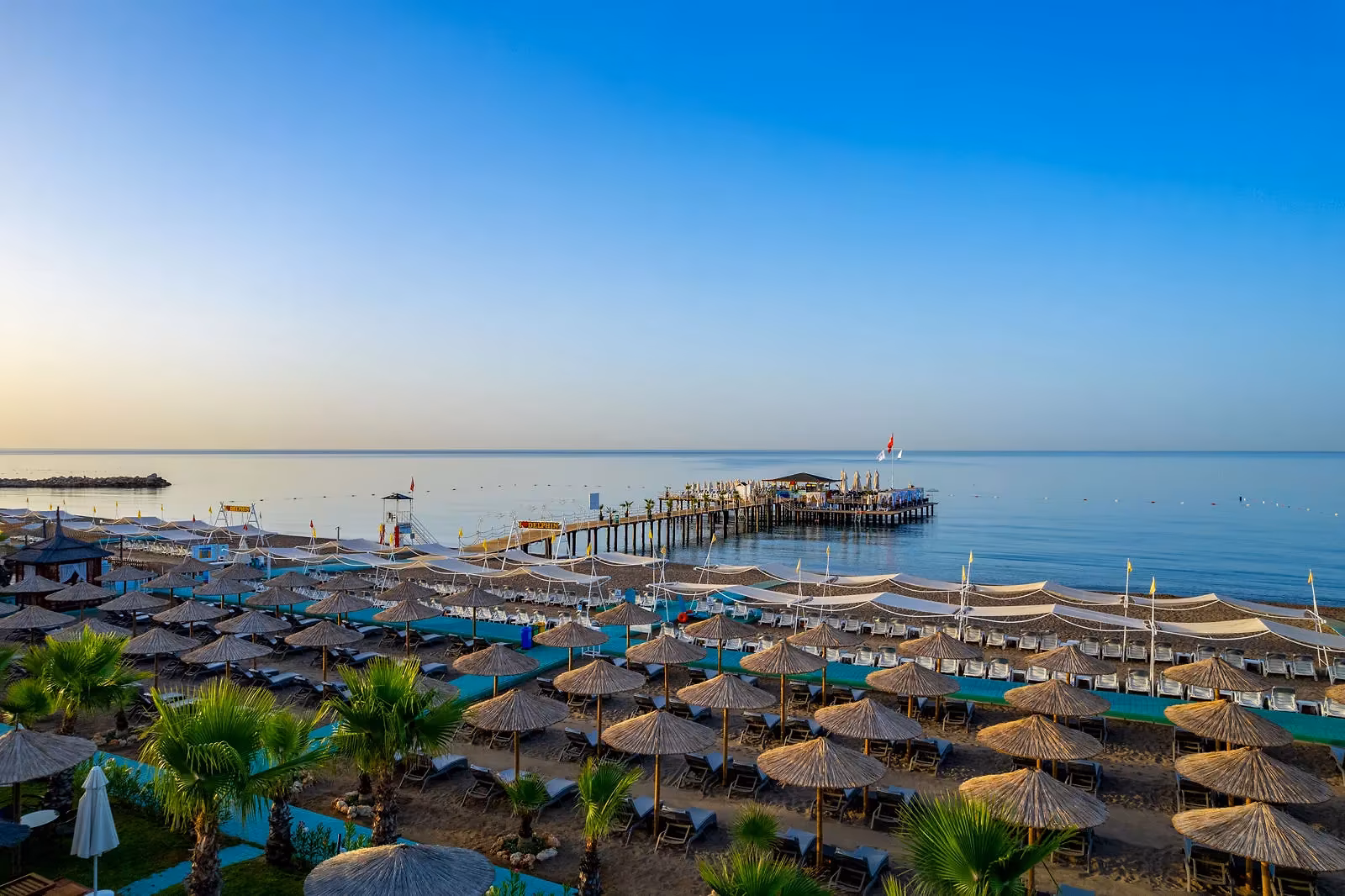 Zandstrand met rieten parasols en ligbedden en de pier in zee bij zonsondergang bij Delphin Be Grand