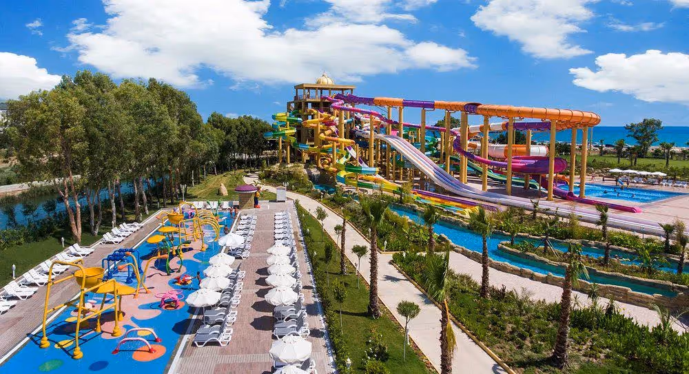 Luchtfoto van een lang zwembad met ligbedden, een kinderspeelbad en de glijbanen erachter bij Delphin BE Grand Resort in Lara